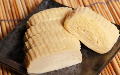 Trzy porcje nigiri tamago, delikatnie zwiniętego omletu jajecznego, ułożone na czarnym talerzu na tle bambusowej maty.