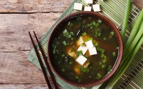 Aksamitne miso z tofu, wodorostami i szczypiorkiem, podane z pałeczkami na drewnianym blacie.