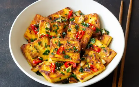 Chrupiące kawałki tofu satay, posypane chili, sezamem i ziołami, podane w białej misce z pałeczkami.