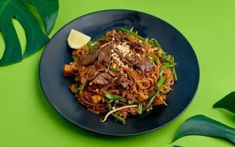 Pyszne pad thai z wołowiną, fasolką szparagową i orzeszkami na czarnym talerzu, ozdobione ćwiartką cytryny i liśćmi monstery.
