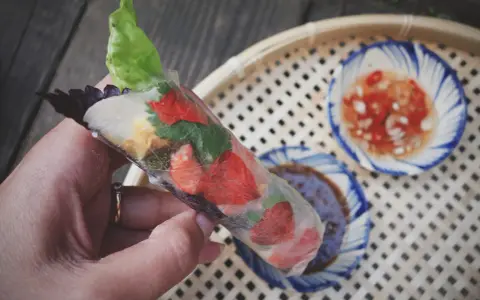 Ręka trzyma świeże rolls z papieru ryżowego z truskawkami i ziołami. Obok miseczki z sosem.