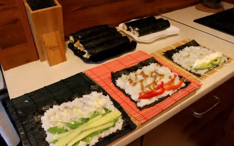 Przygotowanie ryżu do sushi. Na matach leżą arkusze nori z ryżem i dodatkami: ogórkiem, awokado, papryką i sosem.