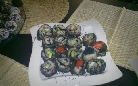 Talerz pełen sushi z sezamem, z widocznymi kawałkami awokado i ikrą. Obok deserki w wiórkach kokosowych.
