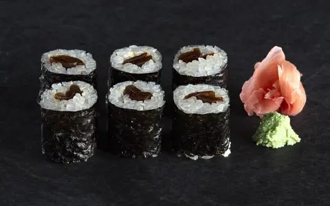 Sześć roladek sushi z ryżem i ciemnym nadzieniem, prawdopodobnie sushi z tykwą. Obok różowy imbir i zielone wasabi.