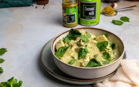Aksamitny kurczak w zielonym curry, udekorowany świeżym szpinakiem i kolendrą. Obok puszka mleka kokosowego i słoiczek pasty curry.