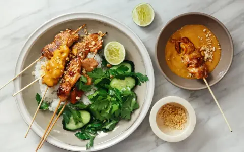 Pyszny kurczak satay na ryżu z orzeszkami ziemnymi, ogórkiem i limonką. Obok miseczka z sosem orzechowym.