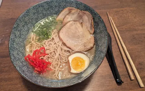 Miseczka z ramen: makaron, plastry mięsa, jajko na pół, zielona cebulka i czerwone przyprawy do ramen. Obok pałeczki.