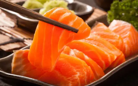 Sashimi co to? Świeże, pomarańczowe kawałki łososia podniesione pałeczkami, obok zielonej pasty wasabi i natki pietruszki.