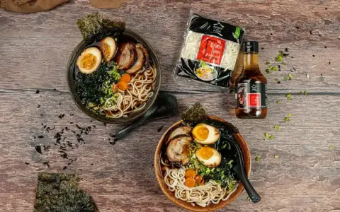 Dwie miski aromatycznego makaronu ramen z sosem sojowym, jajkiem, mięsem i wodorostami. Obok opakowanie makaronu i butelka sosu.