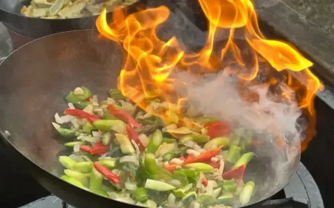 Warzywa w woku płoną w ogniu, tworząc aromatyczny stir fry. W tle widać drugie danie.