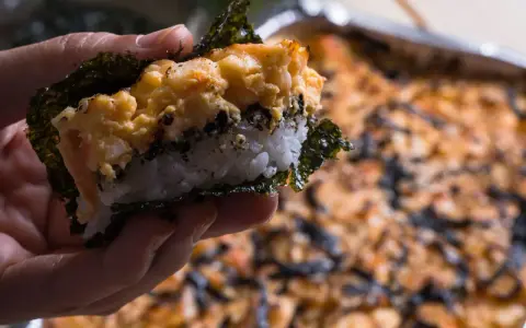 Dłoń trzyma kawałek sushi bake z ryżem, nori i zapiekanym farszem.