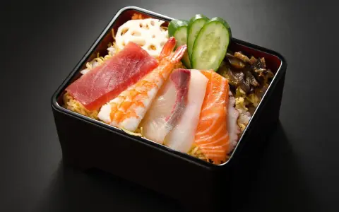 Chirashi zushi: kolorowe plastry ryb (tuńczyk, łosoś, biała ryba), krewetka, plasterki ogórka i lotosu na ryżu.