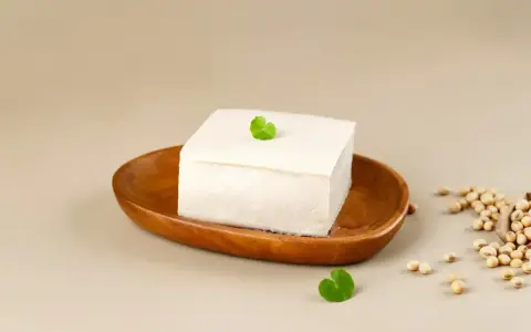 Kostka białego tofu na drewnianej miseczce, ozdobiona listkiem. Obok garść ziaren soi. To właśnie co to tofu – produkt z soi.