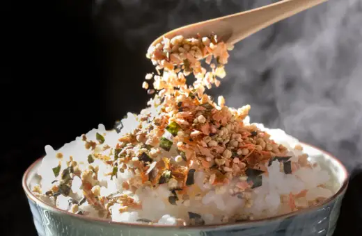 Łyżka sypie na parujący ryż kolorowy furikake. To japońska posypka, która dodaje smaku.