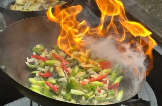 Warzywa w woku płoną w ogniu, tworząc aromatyczny stir fry. W tle widać drugie danie.