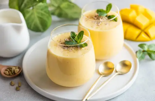 Dwa pyszne mango lassi z pulpy, udekorowane miętą i kardamonem, obok świeżego mango.