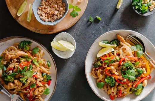 Dwa talerze z prostym pad thai z kurczakiem, brokułami, papryką i orzeszkami ziemnymi, podane z ćwiartkami limonki.