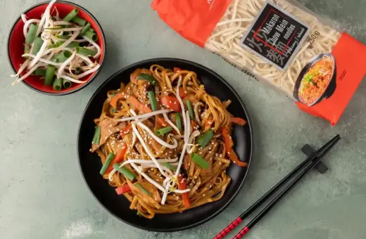 Talerz z makaronem chow mein, kiełkami fasoli mung i szczypiorkiem. Obok paczka makaronu i miseczka z dodatkami.