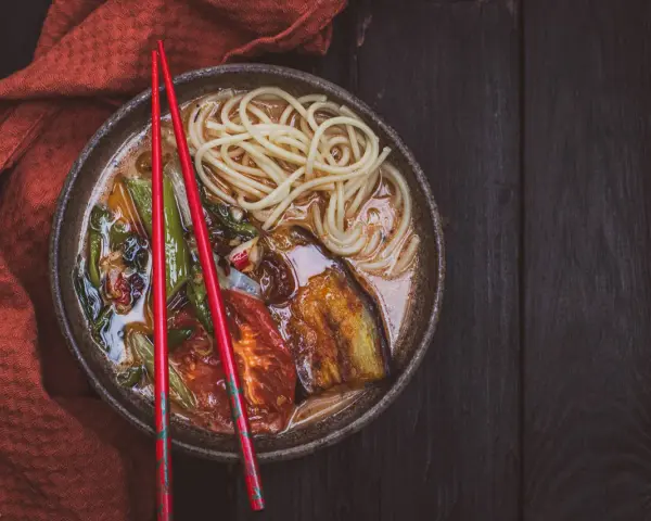 A miseczce z aromatycznym ramen wegetariańskim, pełnej makaronu, grillowanego bakłażana i pomidorów, spoczywają czerwone pałeczki.