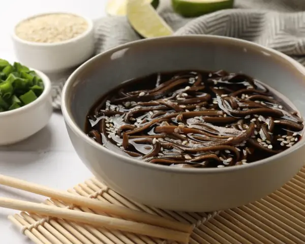 Miseczka z makaronem soba w sosie, posypana sezamem. Obok miseczki z posiekanym szczypiorkiem i sezamem. Zdrowy posiłek, mało makaron soba kcal.