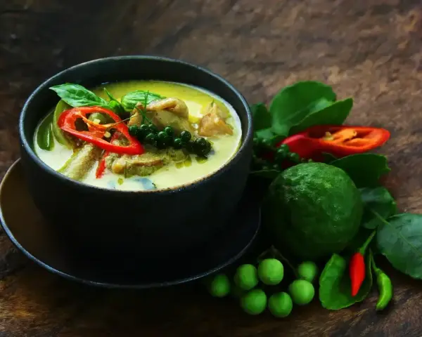 Aromaticzne zielone curry z kawałkami mięsa, papryczką chili, zielonym pieprzem i liśćmi bazylii. Obok miski świeże składniki: limonka kaffir, pomidorki i papryczki chili.