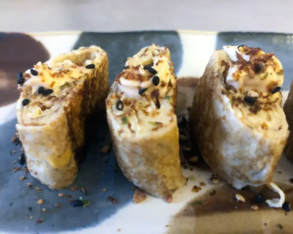 Trzy kawałki sushi w omlecie, polane majonezem i posypane czarnym sezamem.