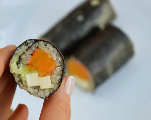 Rolka sushi z tofu, dynią i awokado, trzymana w dłoni.