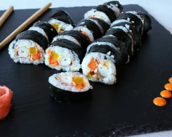 Roladki sushi z surimi, ryżem i warzywami na czarnej desce, obok pałeczki i różowy imbir.