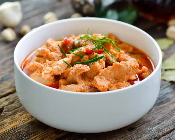 Pyszne wieprzowina w sosie curry, udekorowana szczypiorkiem i papryczką chili, podana w białej misce na drewnianym stole.