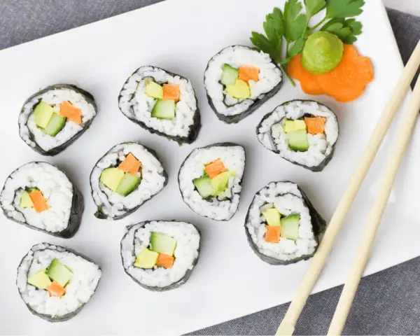 Talerz z rolkami sushi z awokado, ogórkiem i marchewką. Obok pałeczki i dekoracja z marchewki z wasabi. Idealna ilość octu do sushi podkreśla smak.