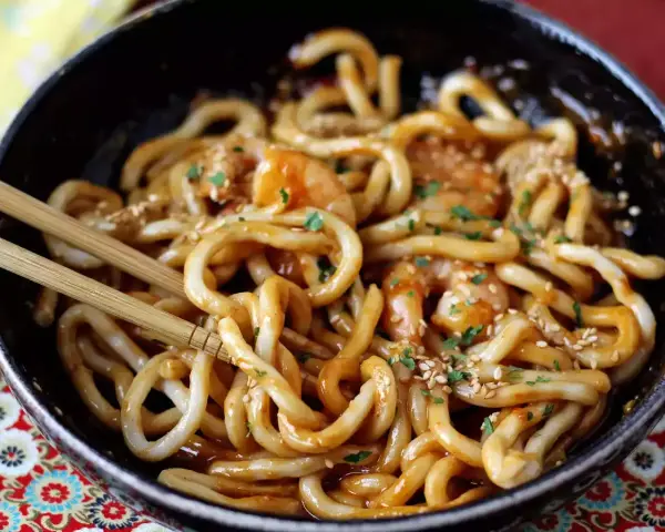 Pyszne udon z krewetkami w sosie, posypane sezamem i natką pietruszki. Danie w czarnej misce, gotowe do jedzenia pałeczkami.