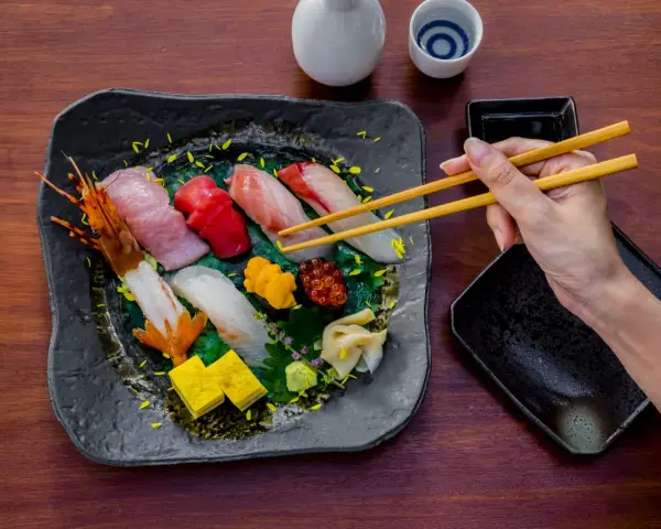 Wykwintne jedzenie w Japonii: ręka z pałeczkami sięga po świeże sushi i sashimi, obok sos i sake.