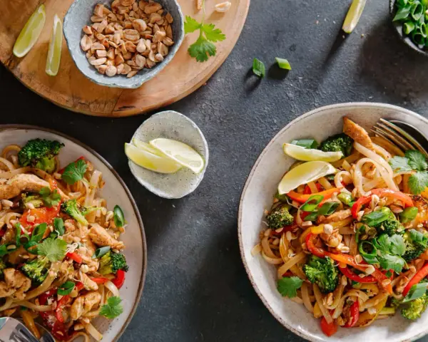 Dwa talerze z prostym pad thai z kurczakiem, brokułami, papryką i orzeszkami ziemnymi, podane z ćwiartkami limonki.