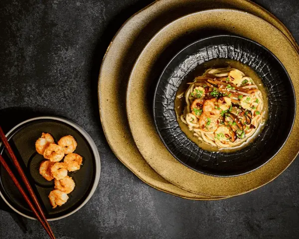 Chintan ramen z krewetkami i warzywami, podany na czarnym talerzu, obok miseczki z dodatkowymi krewetkami i pałeczkami.