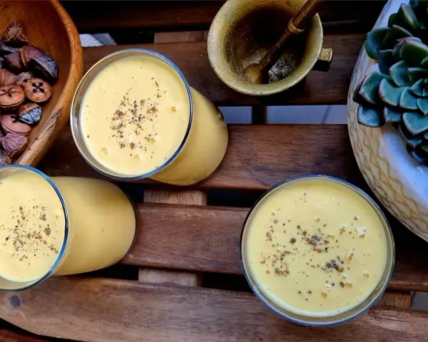 Trzy szklanki kremowego mango lassi, posypane przyprawami, obok miski z suszonymi owocami i sukulentu.