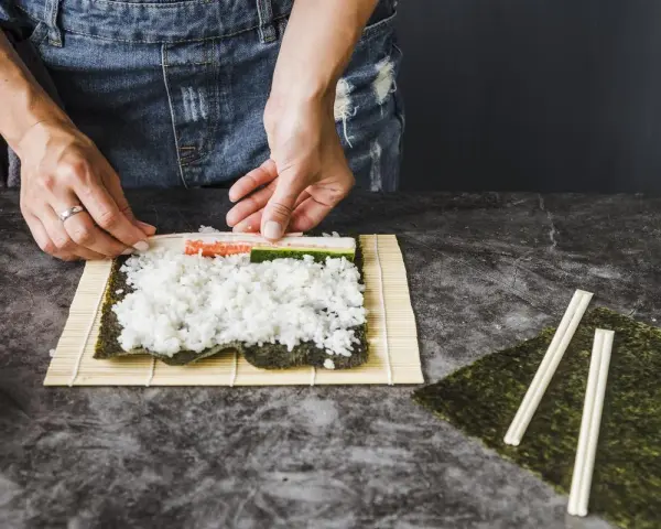 Ręce układają składniki do zwijania sushi na macie bambusowej. Ryż, paluszki krabowe i ogórek czekają na zawinięcie.