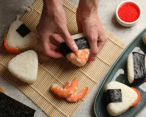 Ręce formują onigiri z krewetkami, tworząc pyszne japońskie jedzenie. Obok miska ryżu, nori i talerz gotowych przysmaków.