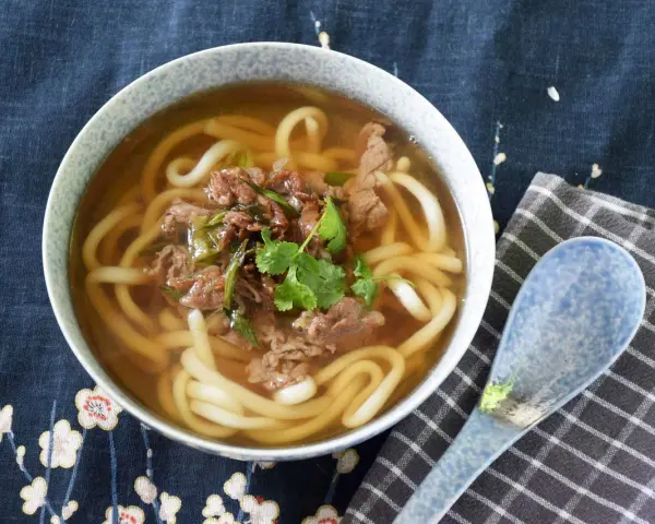 Gorąca zupa udon z wołowiną i szczypiorkiem, ozdobiona kolendrą, w misce na tle z motywem kwiatowym. Obok leży niebieska łyżka.