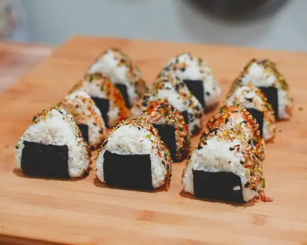 Rząd trójkątnych onigiri z kurczakiem, posypanych sezamem i przyprawami, ułożonych na drewnianej desce.