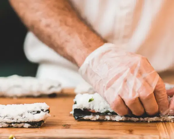 Ręce w rękawiczkach zwijają sushi na desce. Dowiedz się, jak zrobić zaprawę do ryżu sushi, by uzyskać idealny smak.