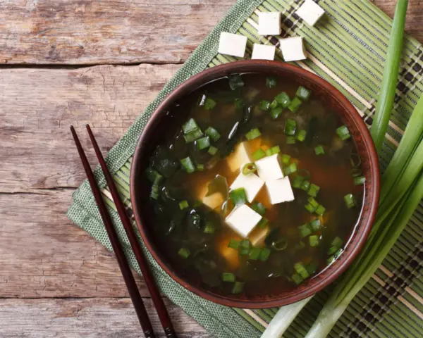 Aksamitne miso z tofu, wodorostami i szczypiorkiem, podane z pałeczkami na drewnianym blacie.