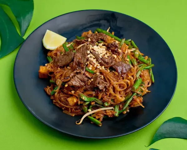 Pyszne pad thai z wołowiną, fasolką szparagową i orzeszkami na czarnym talerzu, ozdobione ćwiartką cytryny i liśćmi monstery.