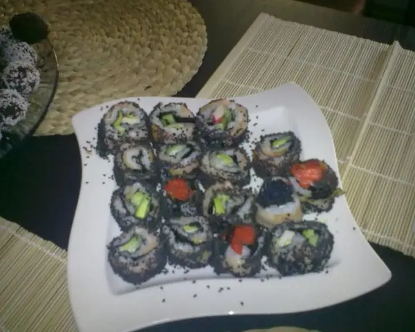 Talerz pełen sushi z sezamem, z widocznymi kawałkami awokado i ikrą. Obok deserki w wiórkach kokosowych.