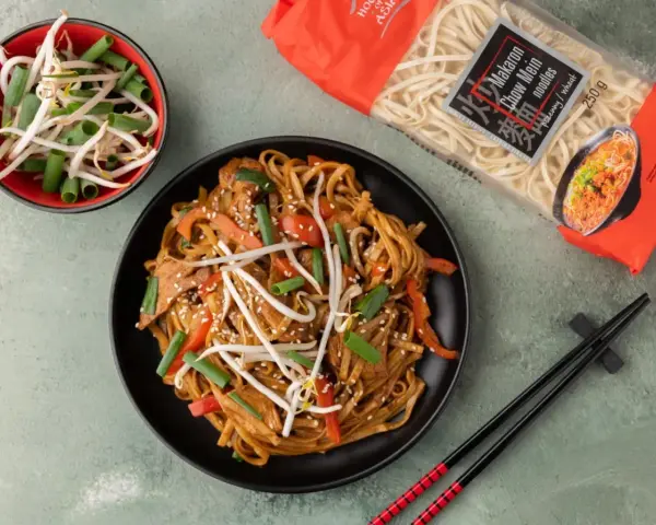 Pyszny makaron sojowy z kurczakiem jak od chińczyka, z kiełkami fasoli mung i szczypiorkiem. Obok opakowanie makaronu chow mein.