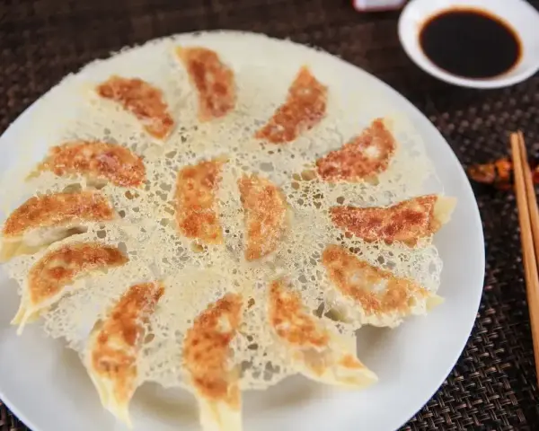 Chrupiące pierożki gyoza ułożone na talerzu, z chrupiącą panierką z ciasta. Obok miseczka sosu sojowego i pałeczki.