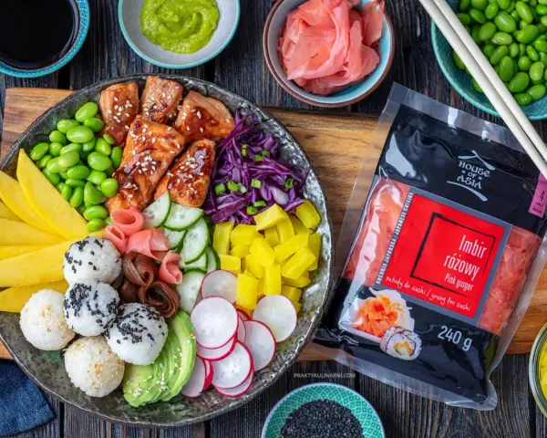 Kolorowy bowl z kawałkami glazurowanego lososia, ryżem, awokado, mango, ogórkiem, rzodkiewką, edamame i marynowanym imbirem. Obok opakowanie różowego imbiru.