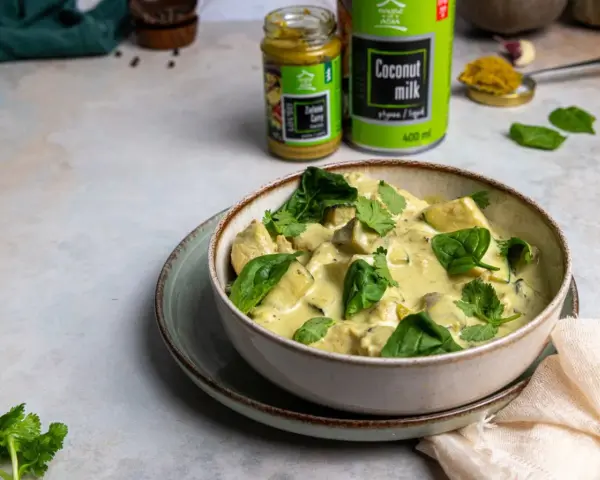 Aksamitny kurczak w zielonym curry, udekorowany świeżym szpinakiem i kolendrą. Obok puszka mleka kokosowego i słoiczek pasty curry.
