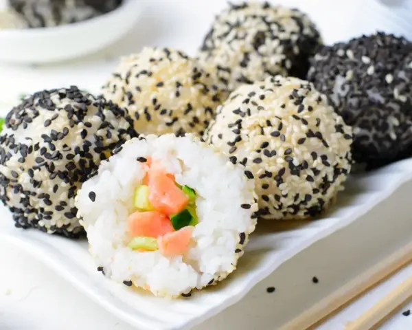 Szybkie sushi w kulkach, obtoczone białym i czarnym sezamem. Jedna kulka jest przekrojona, ukazując łososia i ogórek.