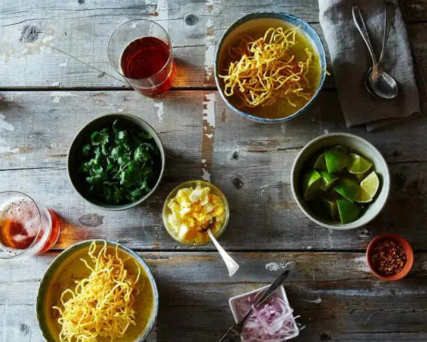 Dwa miseczki khao soi, świeża kolendra, limonki, jajko na twardo, czerwona cebula i płatki chili na drewnianym stole.