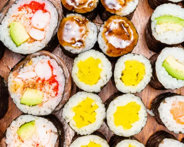 Różnorodne rolki sushi: grubsze futomaki z krewetkami i awokado, cieńsze hosomaki z łososiem, awokado, żółtą rzodkwią i polane sosem.